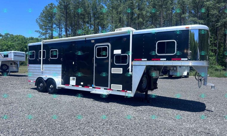 Black and silver horse trailer on a gravel lot with a wooded backdrop and blue sky behind it.