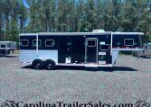 Black horse trailer with silver lower trim parked on a gravel lot, trees in the background.