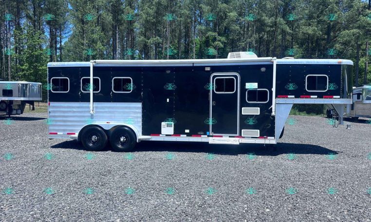 Black horse trailer with silver lower trim parked on a gravel lot, trees in the background.