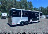 Long black and silver horse trailer in a gravel lot with pine trees and a blue sky behind.