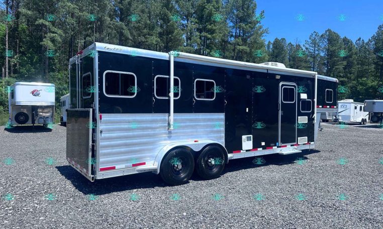 Long black and silver horse trailer in a gravel lot with pine trees and a blue sky behind.