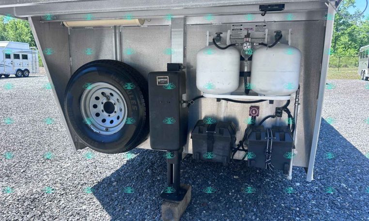 Front storage bay of a travel trailer with a spare tire, two propane tanks, and battery boxes; visible gas lines and gauges.