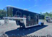 Black gooseneck tandem-axle trailer with chrome trim and side windows, parked on a gravel lot under a blue sky with trees in the background. The trailer has a raised cab area and exterior steps on the side.
