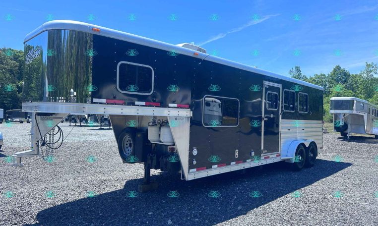 Black gooseneck tandem-axle trailer with chrome trim and side windows, parked on a gravel lot under a blue sky with trees in the background. The trailer has a raised cab area and exterior steps on the side.