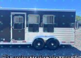 Side view of a dark-colored horse trailer with two barred windows and aluminum trim, resting on gravel with tandem wheels visible