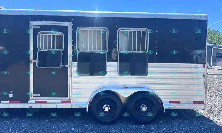 Side view of a dark-colored horse trailer with two barred windows and aluminum trim, resting on gravel with tandem wheels visible