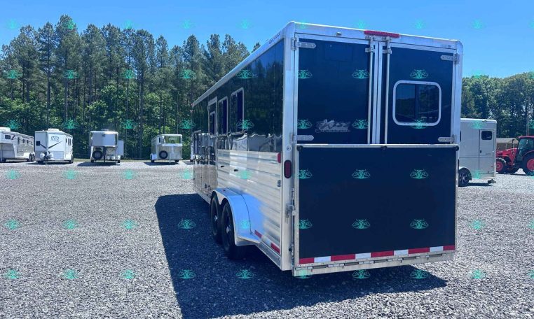 Rear view of a large blue and silver enclosed trailer in a gravel lot, with other trailers and tall trees in the background. Watermark text CarolinaTrailerSales.com across the bottom.