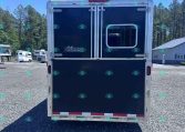 Rear view of a dark blue horse trailer with a silver frame parked on a gravel lot, with other trailers and pine trees in the background.