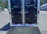 Rear view of a dark blue cargo trailer with large double doors, a lowered ramp, and a small window on the right door in a sunny gravel lot.
