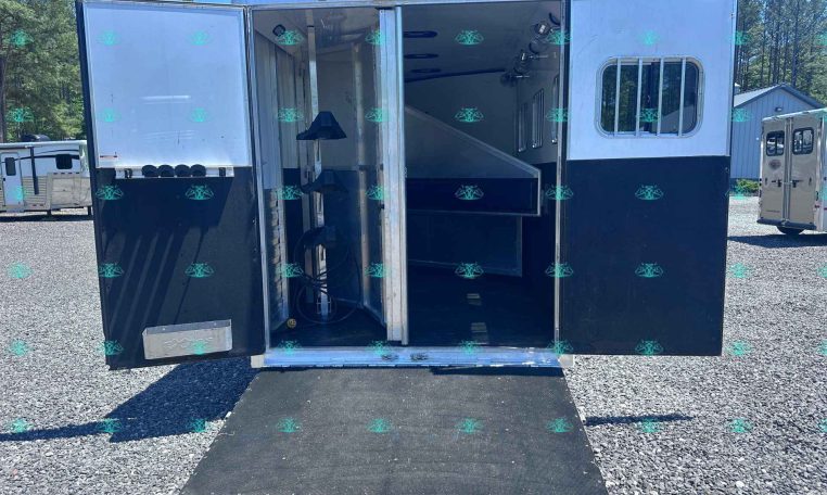 Open horse trailer with ramp down, showing interior stalls and tack area against a gravel lot and blue sky.