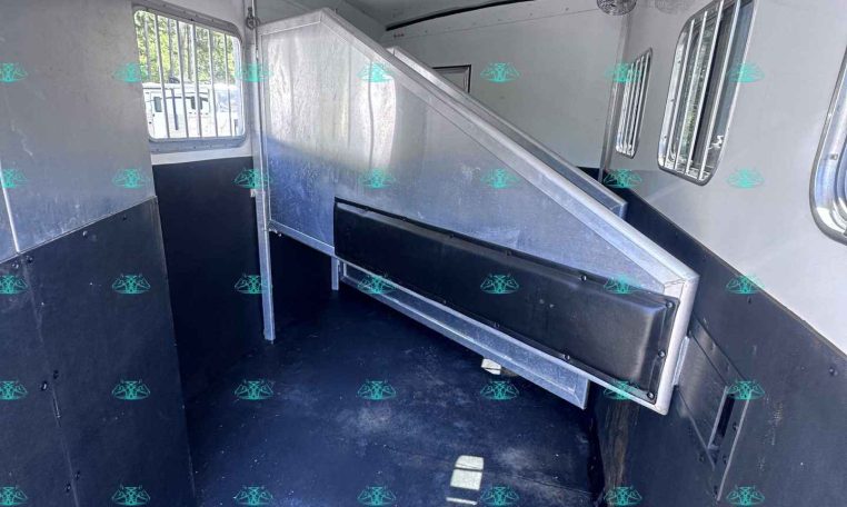 Interior of a metal horse trailer with a divider and barred windows, showing the parking ramp built into the stall area, and ceiling vents/speakers above.