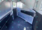 Interior of a horse trailer with metal dividing stalls, black padding, and side windows inside a white/gray aluminum body.