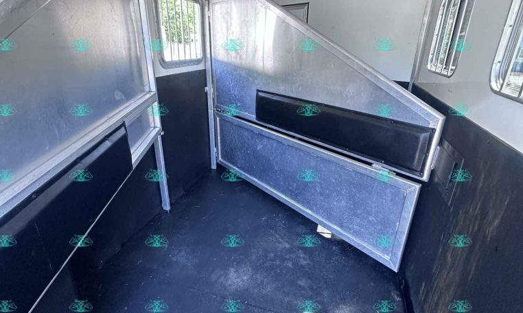 Interior of a horse trailer with metal dividing stalls, black padding, and side windows inside a white/gray aluminum body.
