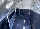 Interior of a metal horse trailer with a fold-down divider on the left, doorway at far end, and windows along the right side.