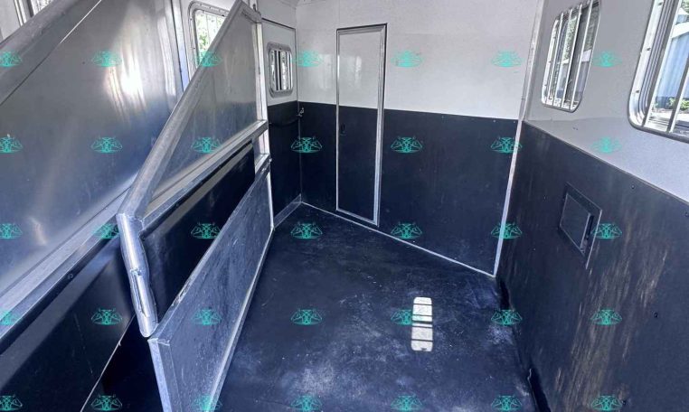 Interior of a metal horse trailer with a fold-down divider on the left, doorway at far end, and windows along the right side.