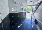 Interior of an empty white horse trailer with black lower panels, side windows with bars, ceiling vents/fans, and an open rear doorway showing a gravel lot outside.