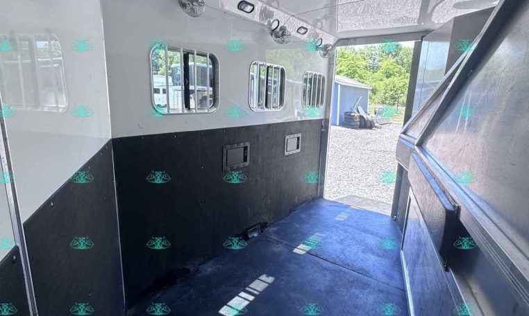 Interior of an empty white horse trailer with black lower panels, side windows with bars, ceiling vents/fans, and an open rear doorway showing a gravel lot outside.