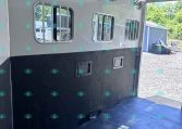 Interior of a white horse trailer with three barred side windows, ceiling vent fans, and a black lower wall; gravel lot visible outside.