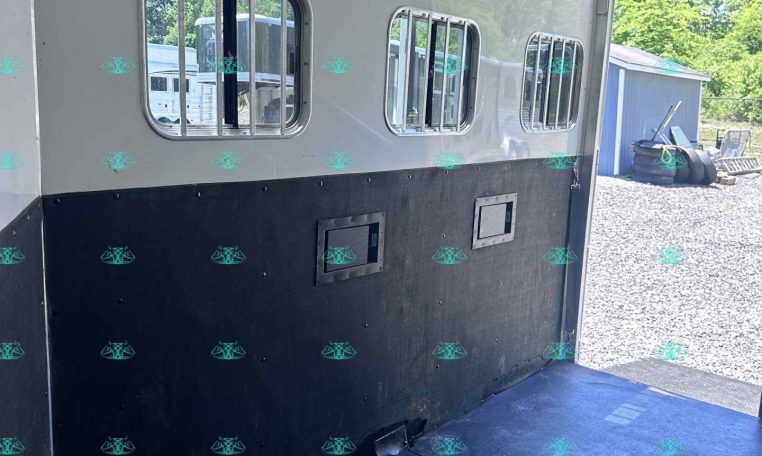 Interior of a white horse trailer with three barred side windows, ceiling vent fans, and a black lower wall; gravel lot visible outside.
