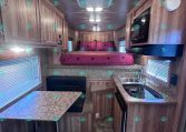 Interior of a camper with a kitchen, corkstone counters, and wooden cabinetry leading to a bed with pink bedding at the rear.