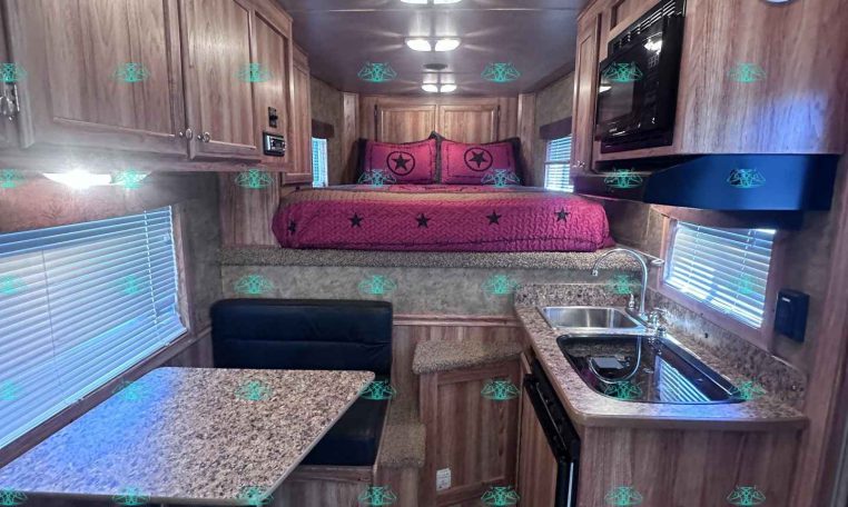 Interior of a camper with a kitchen, corkstone counters, and wooden cabinetry leading to a bed with pink bedding at the rear.