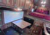Interior of a wooden RV with upper cabinets, a pink bed with star pillows along the wall, and a tiled kitchenette countertop nearby.