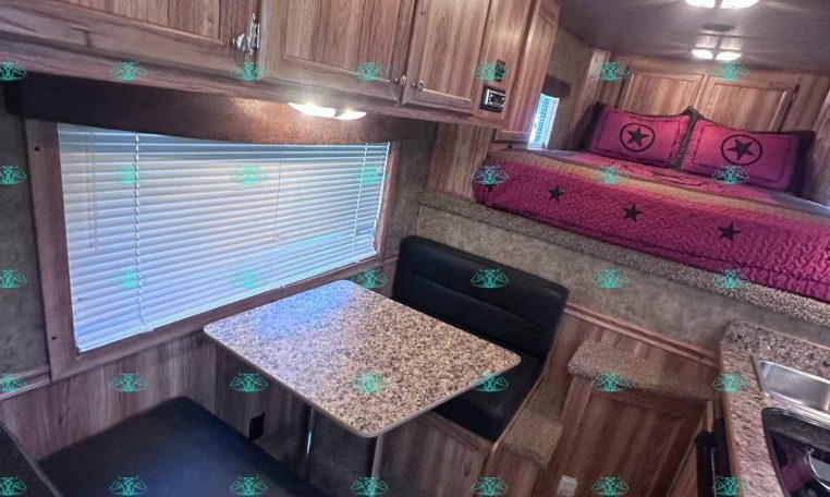 Interior of a wooden RV with upper cabinets, a pink bed with star pillows along the wall, and a tiled kitchenette countertop nearby.