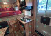 RV interior with a bed and pink quilt, built-in wood cabinets, microwave, and small kitchen with stove and sink.