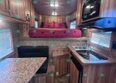 A compact RV kitchen with granite counters, wooden cabinets, stainless sink, and a cooktop on the right, leading to a raised bedroom area with pink bedding and star pillows at the back.