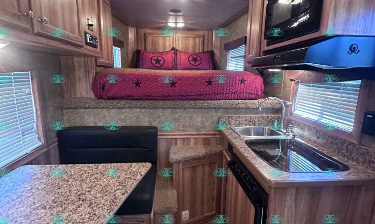 A compact RV kitchen with granite counters, wooden cabinets, stainless sink, and a cooktop on the right, leading to a raised bedroom area with pink bedding and star pillows at the back.