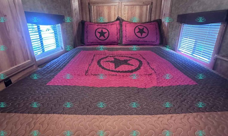Interior of a cozy trailer bedroom with a pink quilt and matching star pillows on a bed against wooden cabinets.