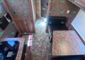 Interior of a camper kitchen with granite countertops, wooden cabinets, and a stove on the left; a small island on the right; doorway to a bathroom in the back.