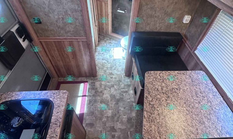 Interior of a camper kitchen with granite countertops, wooden cabinets, and a stove on the left; a small island on the right; doorway to a bathroom in the back.
