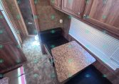 Interior of a wood-paneled RV kitchen with upper cabinets, a speckled countertop, and a black cushioned dinette seat by a window with blinds