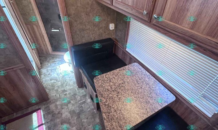 Interior of a wood-paneled RV kitchen with upper cabinets, a speckled countertop, and a black cushioned dinette seat by a window with blinds