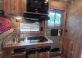 Compact RV kitchen with wooden cabinets, microwave, stove, and double sink under a window with blinds