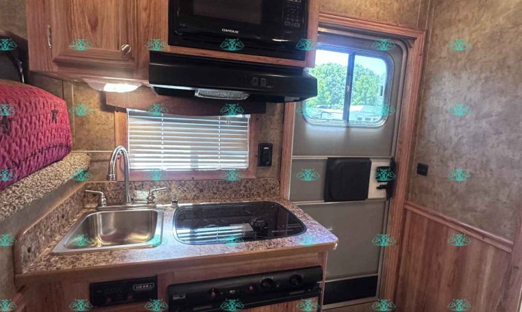 Compact RV kitchen with wooden cabinets, microwave, stove, and double sink under a window with blinds