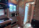 RV kitchen interior with wooden cabinets, granite countertop, stove, and microwave; windowed door to outside visible on left and hallway to a bathroom ahead.
