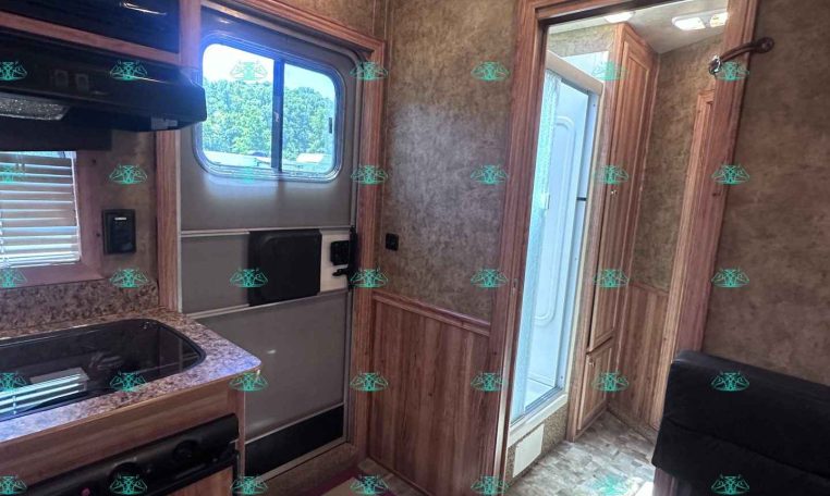 RV kitchen interior with wooden cabinets, granite countertop, stove, and microwave; windowed door to outside visible on left and hallway to a bathroom ahead.
