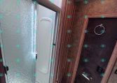 Compact RV bathroom with a frosted glass shower door, textured shower wall, and wood-grain cabinetry above. A circular towel ring sits on a dark door nearby with ceiling lights overhead.