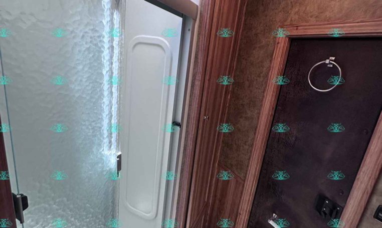 Compact RV bathroom with a frosted glass shower door, textured shower wall, and wood-grain cabinetry above. A circular towel ring sits on a dark door nearby with ceiling lights overhead.