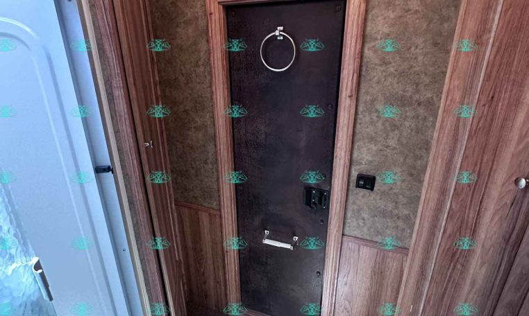 RV bathroom with a frosted shower door on the left, dark central access door with a circular towel ring, and wood paneling.