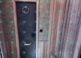 RV bathroom interior with brown wood cabinets, beige textured walls, and a black metal access door featuring a circular towel ring above the latch. A blue toilet seat is in the foreground.