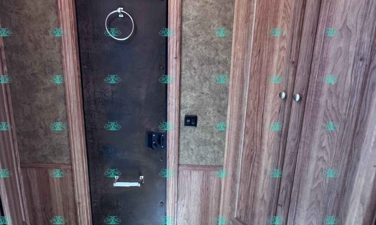 RV bathroom interior with brown wood cabinets, beige textured walls, and a black metal access door featuring a circular towel ring above the latch. A blue toilet seat is in the foreground.
