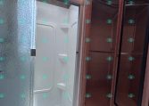 Interior of a travel trailer bathroom showing a frosted glass shower door, white shower stall, skylight above, and wood-paneled cabinet with a mirrored door on the right.
