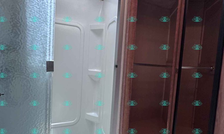 Interior of a travel trailer bathroom showing a frosted glass shower door, white shower stall, skylight above, and wood-paneled cabinet with a mirrored door on the right.