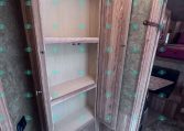 Open wooden cabinet with three shelves inside a trailer living area; beige interior, adjacent door, and a bed with red bedding visible in the background.