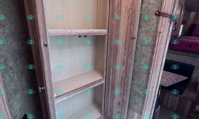 Open wooden cabinet with three shelves inside a trailer living area; beige interior, adjacent door, and a bed with red bedding visible in the background.