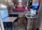 Interior of a compact RV showing a kitchen on the right with sink, stove, microwave, and overhead cabinets, plus a pink-bedding sleeping area at the rear.