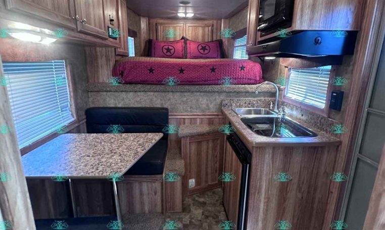 Interior of a compact RV showing a kitchen on the right with sink, stove, microwave, and overhead cabinets, plus a pink-bedding sleeping area at the rear.